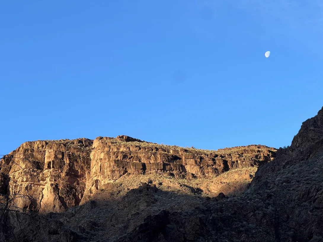 View of a canyon wall with moon above in a clear blue sky